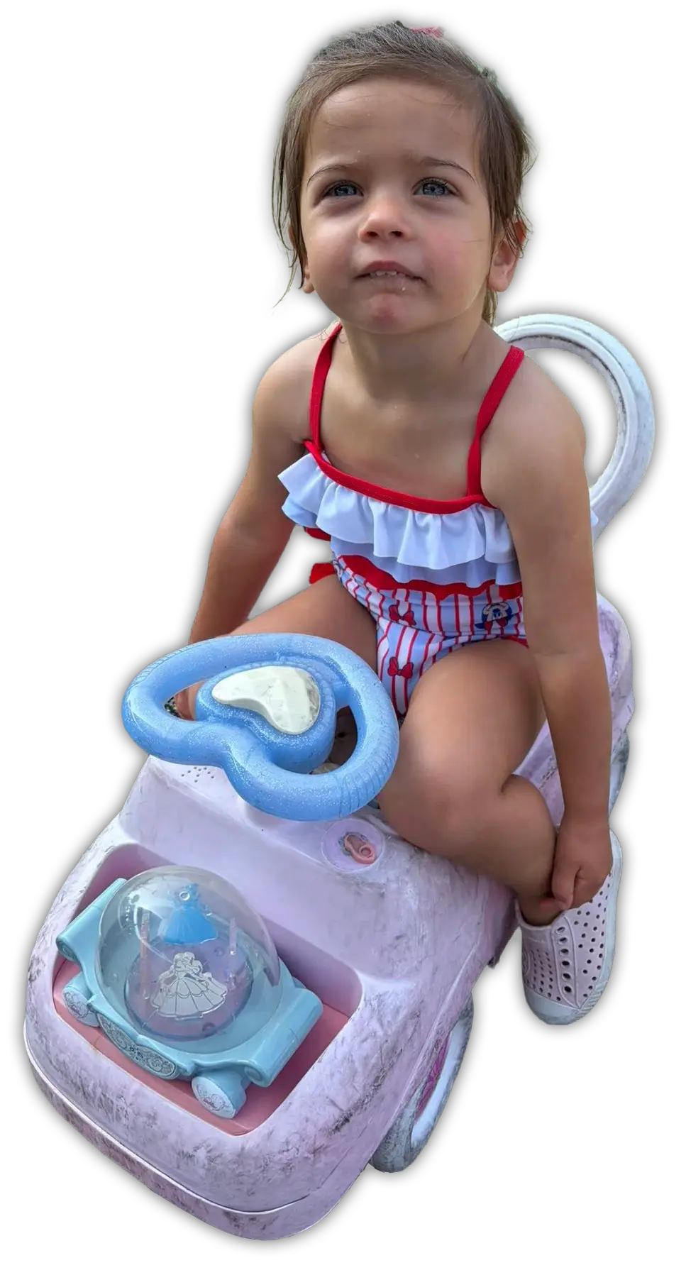 A young girl with blue eyes, wearing a red and white ruffled swimsuit, sits on a worn pink ride-on toy car with a blue ste...