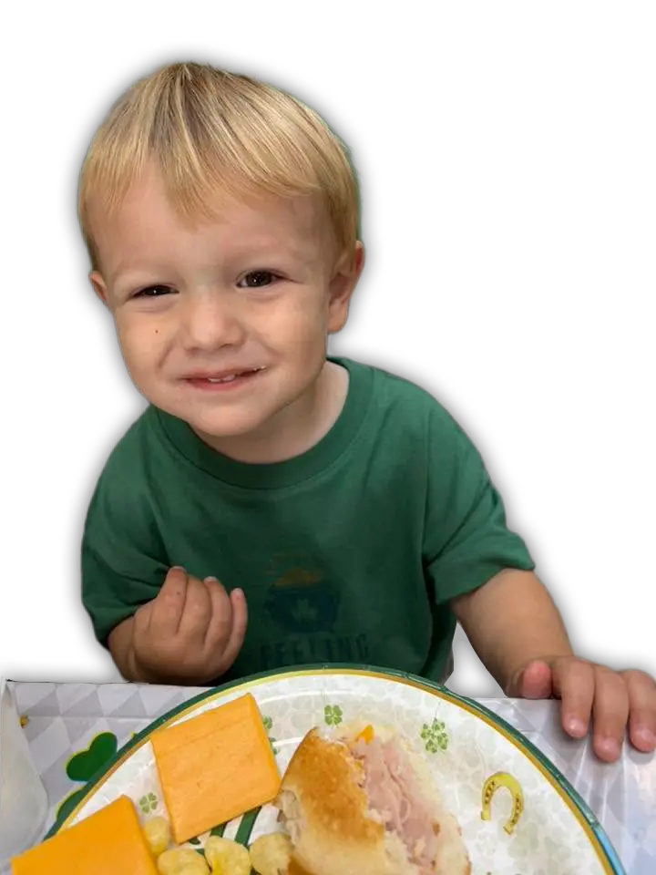 A smiling young blonde boy in a green t-shirt sits at a table with a St. Patrick's Day themed plate of cheese and a sandwi...