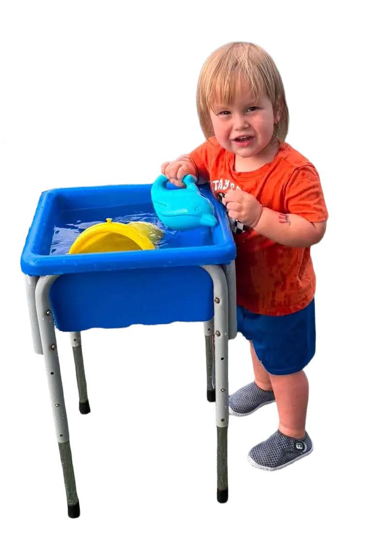 A happy toddler with blonde hair playing with a blue water table and a dolphin toy, wearing a wet orange shirt and blue sh...
